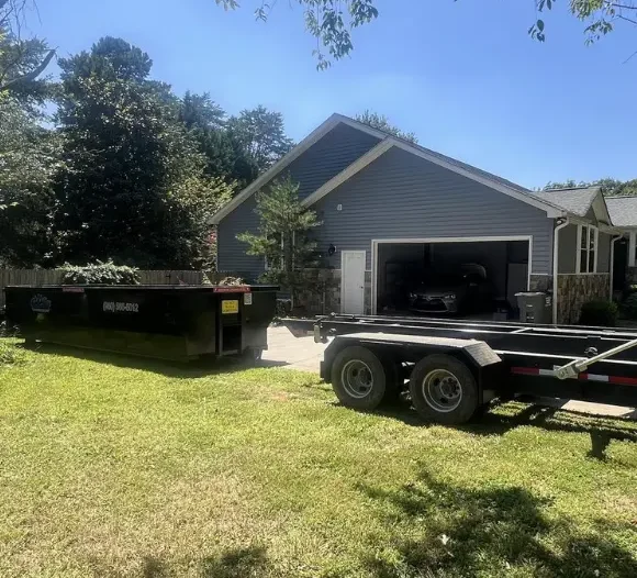 A gray house with an open garage and a black car inside is seen on a sunny day. A large black dumpster and an empty black trailer sit on the driveway.